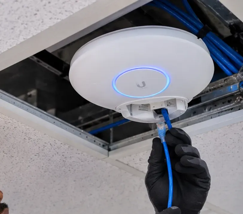 Ceiling access point and its Cat6 feed — Midtown Manhattan White round ceiling access point lowered from an open ceiling tile while a gloved hand holds the blue cable plugged into its base — access point link repair in Midtown Manhattan