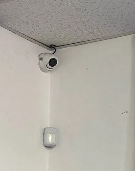 Corner dome camera with cable loop at the ceiling — Mott Haven Cat6 repair reference Reference photo: white dome camera in a room corner with a short black cable loop running up into the ceiling edge, a wall sensor mounted below — exposed cable entry of the kind we re-terminate in Mott Haven, Bronx