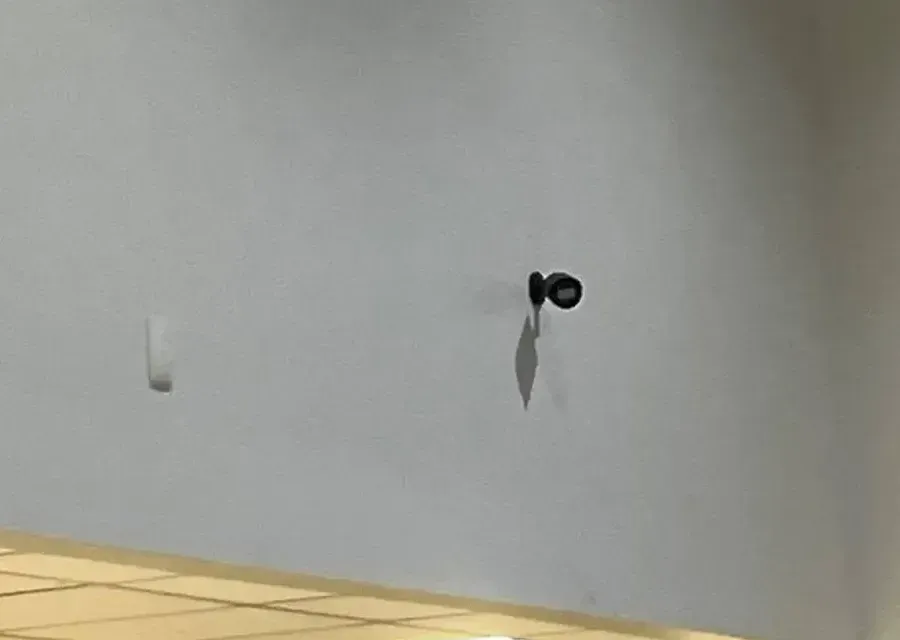 Small camera high on a wall — Mott Haven Cat6 repair reference Reference photo: small black camera on a short arm high on a pale wall, casting a long shadow, beside a white wall box, above the edge of a lowered ceiling — surface-run camera link of the kind we repair in Mott Haven, Bronx