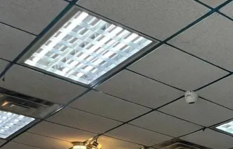 Small dome beside a lay-in light fixture — Cat6 repair reference, Mott Haven Reference photo: small white dome camera on a dark-gridded drop ceiling beside a lay-in light fixture and an air vent — ceiling device link of the kind we test in Mott Haven, Bronx