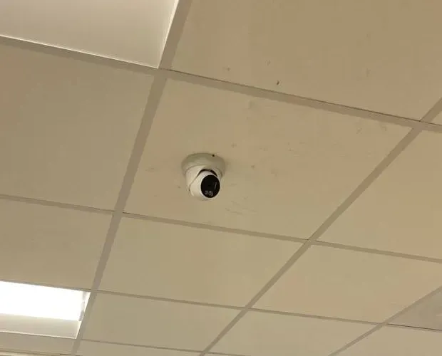 Turret camera in a drop-ceiling tile — Cat6 repair reference, Mott Haven Reference photo: white turret camera mounted in the middle of a drop-ceiling tile, with a light panel in the corner of the grid — ceiling-fed camera link of the type we repair in Mott Haven, Bronx