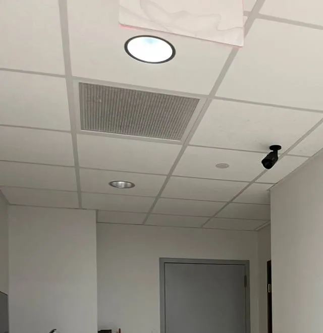 Black bullet camera on a ceiling grid looking down a corridor toward a grey door, with a vent and round light in the ceiling — reference photo from another AESS job, Cat6 repair page for Tottenville, Staten Island