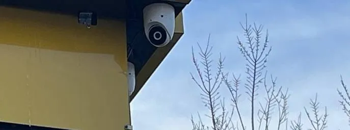White dome camera mounted under the edge of a yellow building fascia against a blue sky and bare branches — reference photo from another AESS job, Cat6 repair page for Tottenville, Staten Island