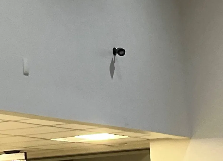 Small black camera high on a pale wall above the edge of a drop ceiling, a white sensor to its left — reference photo from another AESS job, Cat6 repair page for Tottenville, Staten Island