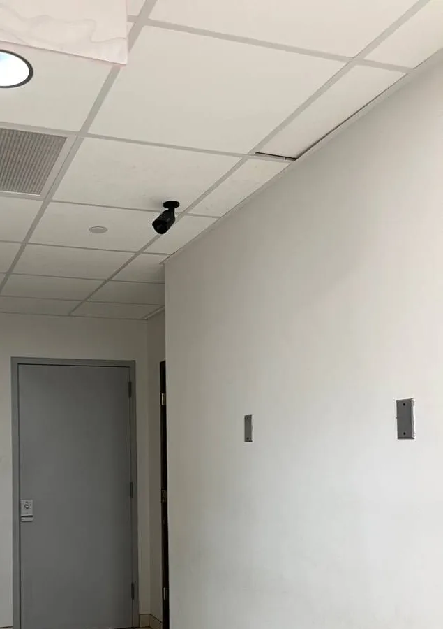 Long Island Dahua camera installation example photo: a black bullet camera mounted at the ceiling corner of a narrow tiled corridor, aimed toward a grey steel door