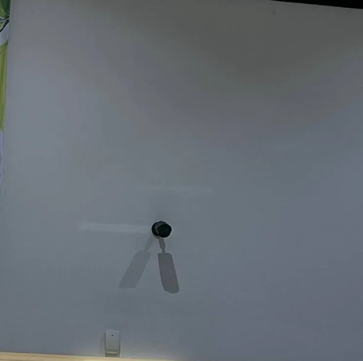 Orange County Dahua wall-mount example: a dark bullet camera on a short arm projecting from a tall pale wall, casting two long shadows, with a motion sensor near the ceiling line below