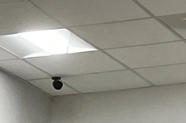 Suffolk County Dahua camera installation example photo: a small black mini dome camera hanging from a white drop-ceiling grid near a square light panel, above a pale wall