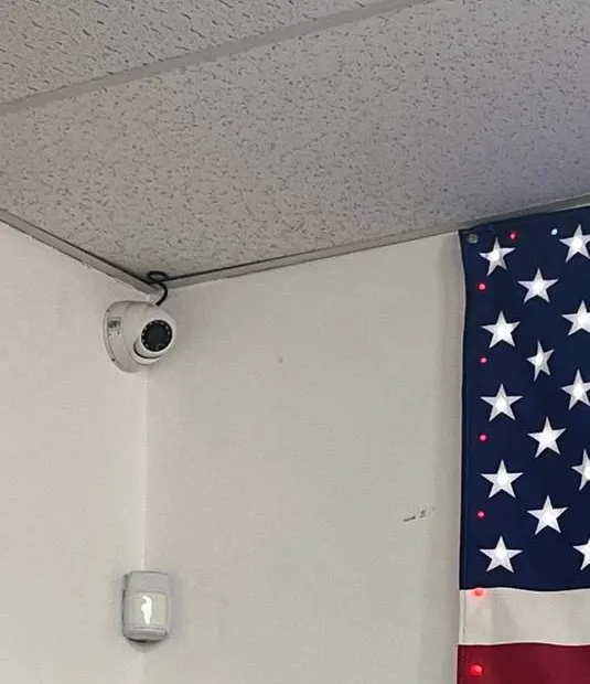 Suffolk County Dahua camera installation example photo: a white turret camera on a short bracket in a ceiling corner above a wall-mounted motion sensor, with an American flag hanging at the right