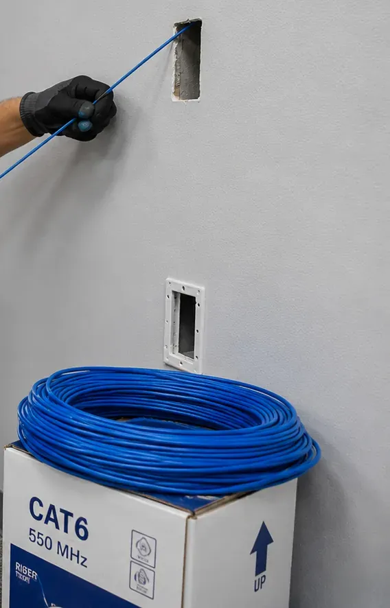 Fish tape into a small wall opening, Charleston Staten Island retrofit route Gloved hand guiding a fish tape into a small opening in a finished wall above a coil of cable on a box, retrofit cable route in Charleston, Staten Island