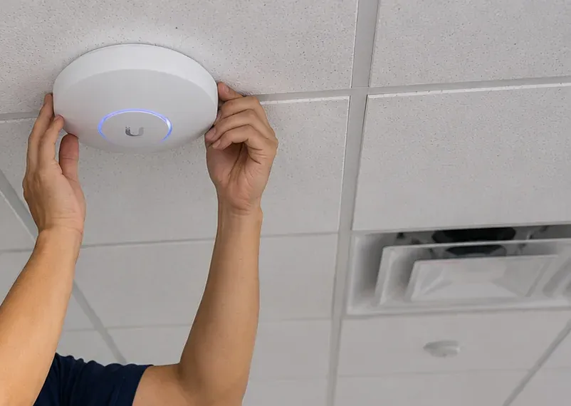 Ceiling access point being fitted to a tile grid, College Point Queens Hands pressing a round white wireless access point onto a ceiling tile, one of the devices fed from fiber optic installation in College Point, Queens
