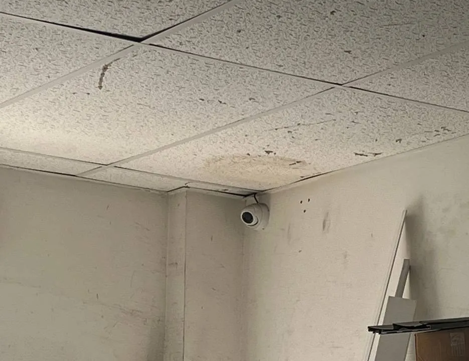 White dome camera in a storeroom corner â Manhattan Hik-Connect support example Manhattan Hik-Connect app support example photo: a small white dome camera mounted high in the corner of a storeroom where a stained drop ceiling meets scuffed white walls