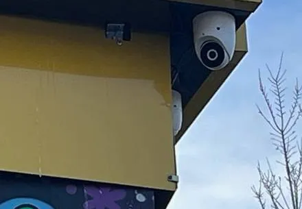 White turret camera under a storefront fascia â Manhattan Hik-Connect support example Manhattan Hik-Connect app support example photo: a white turret camera fixed under the corner of a yellow storefront fascia against a blue sky, with a second camera partly visible below it
