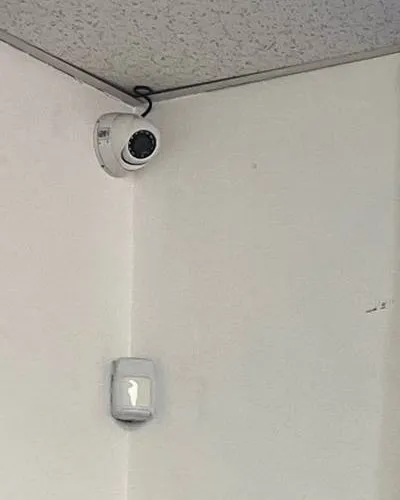 Dome camera and motion sensor in a ceiling corner, example photo for Putnam County office entries Putnam County intercom repair example photo: a small white dome camera at the top of a room corner under a textured ceiling, with a white wall-mounted motion sensor below it