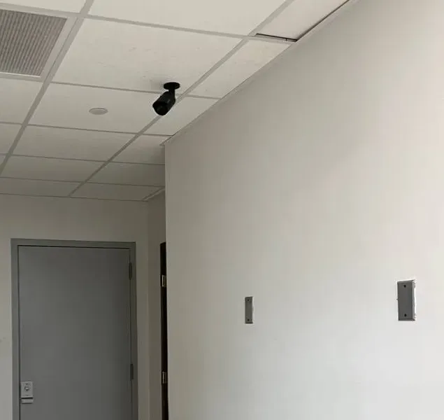 Corridor to a grey door with a ceiling camera and wall plates — example photo for Staten Island service entrances Staten Island intercom repair example photo: a black bullet camera on a tiled ceiling facing a grey door with a keypad lock at the end of a white corridor, with two small metal plates on the wall