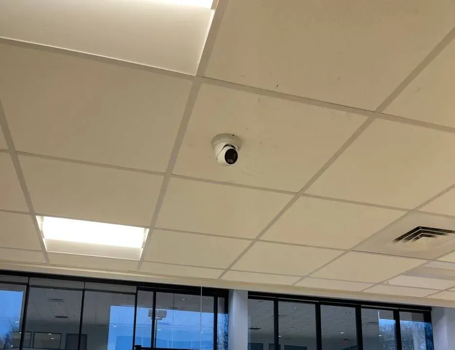 Dome camera on a tiled ceiling above a glass entrance — example photo for Staten Island commercial entries Staten Island intercom repair example photo: a white dome camera on a tiled drop ceiling between two light panels and an air vent, above a wall of glass panes and doors