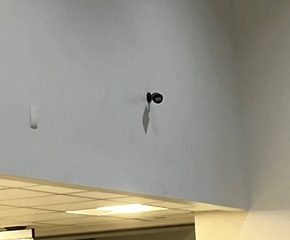 Bullet camera mounted high on a wall above a dropped ceiling β example for Queens booth and key-room coverage Queens parking lot camera installation example photo: a small black bullet camera mounted high on a pale wall above a dropped ceiling