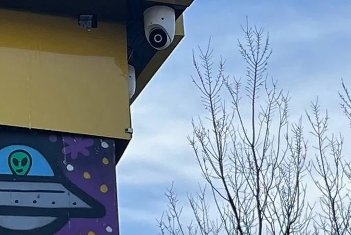 Dome camera under a storefront fascia β example of sheltered exterior mounting for Queens lots Queens parking lot camera example photo: a white dome camera under a yellow storefront fascia beside a painted mural, with a bare tree against the sky