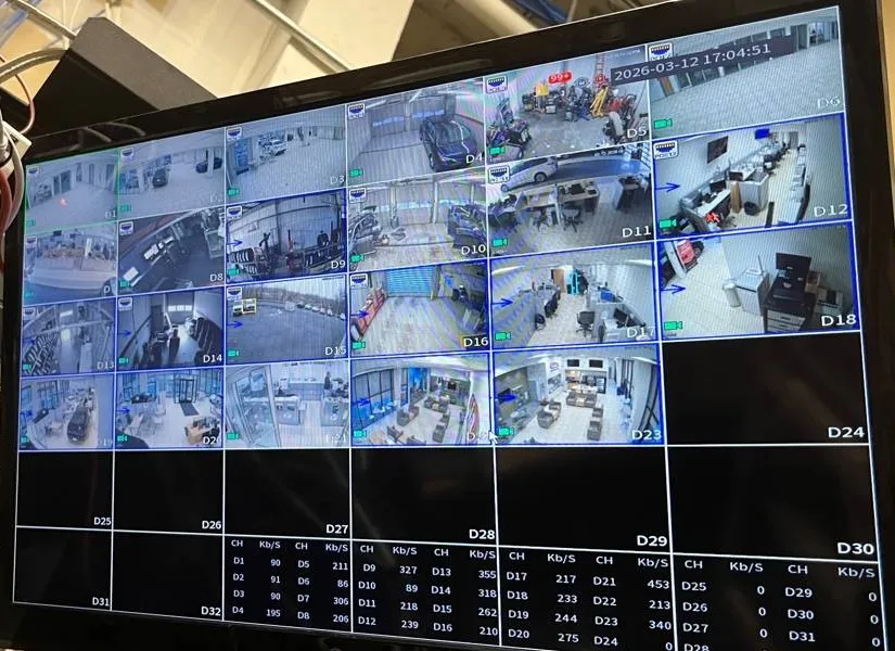 Recorder monitor with a grid of dealership camera views β example for Queens parking lot camera installation Parking lot camera installation Queens example photo: a recorder monitor showing a grid of camera tiles from a car dealership, including service bays, offices and parked cars, with a per-channel bitrate table below