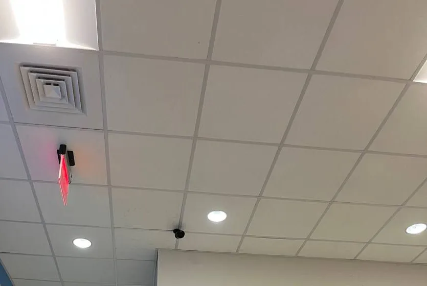 Small ceiling camera near an exit sign β example for Queens garage stair and pedestrian-door coverage Queens parking lot camera installation example photo: a small black camera on a white tiled ceiling near an illuminated exit sign and air vents