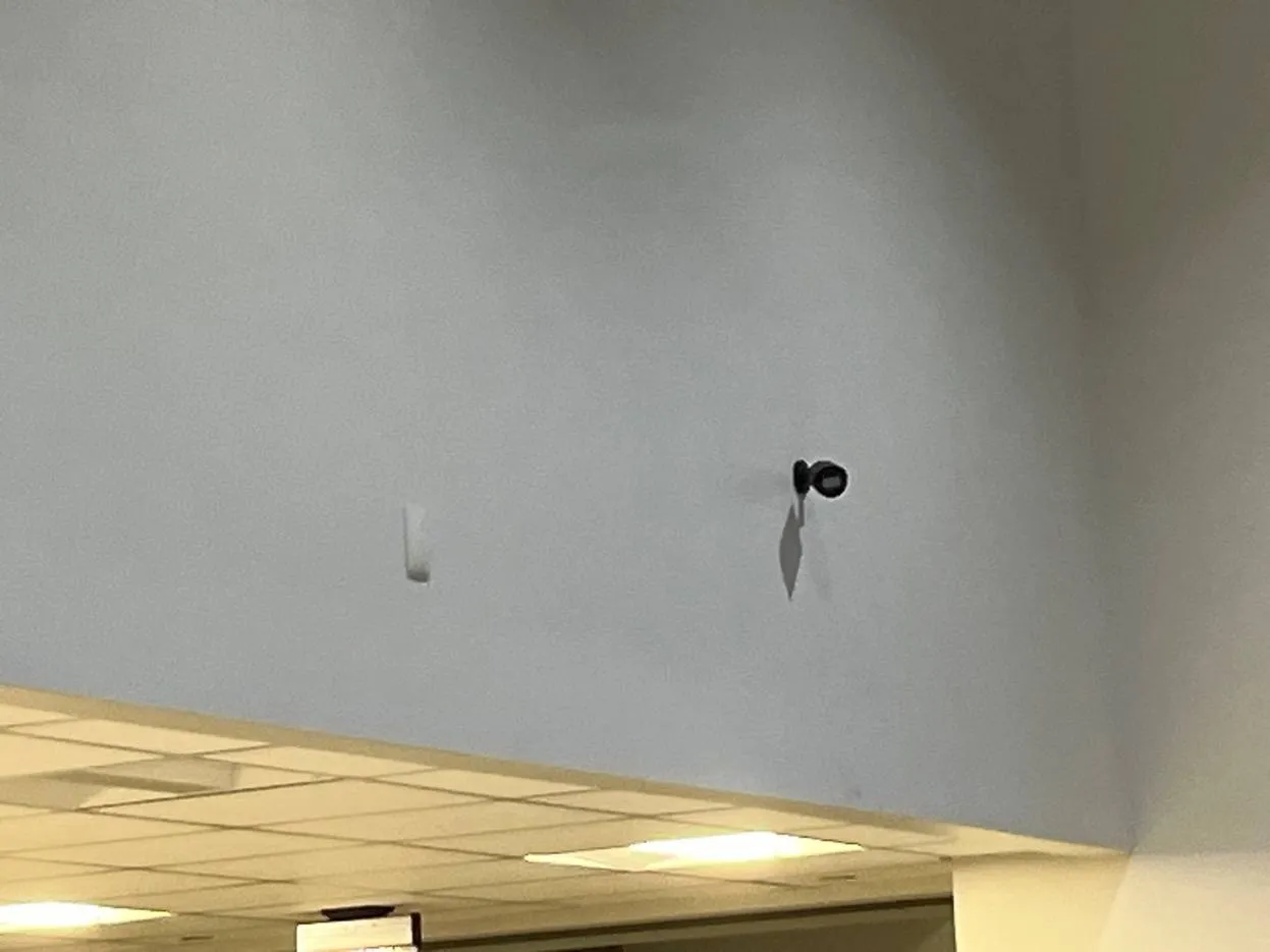 Black camera high on a grey wall above a drop ceiling — example photo for Long Island school gyms and atriums Long Island school security camera installation example photo: a small black camera mounted high on a pale grey wall above a drop ceiling, casting a shadow, with a white wall sensor to its left