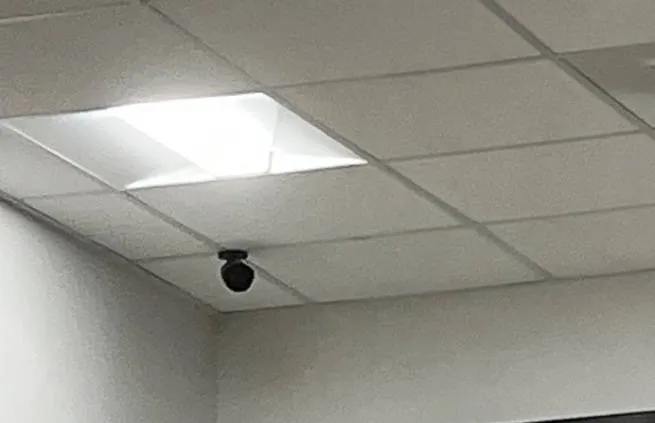 Small black dome on ceiling tiles at the top of a tall wall — example photo for Long Island school lobbies Long Island school security camera installation example photo: a small black dome camera on lay-in ceiling tiles at the top corner of a tall white wall, with a light panel to the left