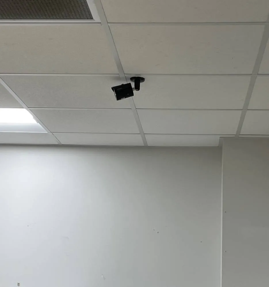 Black bullet camera on a ceiling mount above a plain wall — example photo for Long Island school corridors Long Island school security camera installation example photo: a small black bullet camera on a short ceiling mount in a white lay-in tile ceiling, aimed down along a plain white wall, with a light panel at the left edge