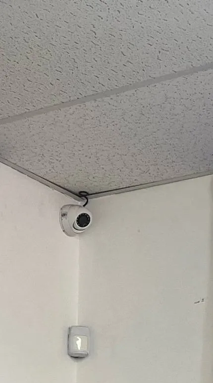 White turret camera in a wall corner above a wall sensor — example photo for Long Island school offices Long Island school security camera installation example photo: a small white turret camera mounted at the top of a wall corner under a textured ceiling, with a white wall sensor below it
