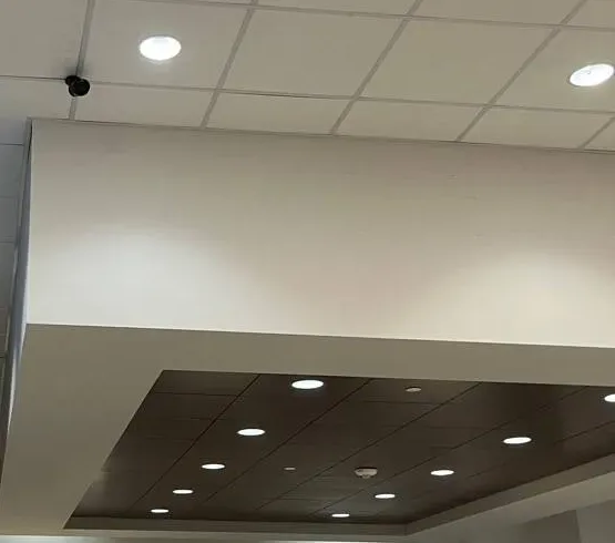 Small camera at a bulkhead edge above a dark coffered ceiling — example photo for Orange County school lobbies Orange County school security camera installation example photo: a small black camera at the edge of a white ceiling bulkhead above a dark recessed coffered ceiling lit by rows of downlights