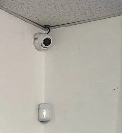 White dome camera above a wall motion sensor — example photo for Orange County schools pairing cameras with alarms Orange County school security camera installation example photo: a white dome camera fixed where a white wall meets a drop ceiling, with a small white wall-mounted motion sensor lower down on the same corner