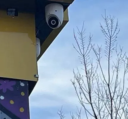 White dome camera under a yellow eave in winter — example photo for Orange County school exteriors Orange County school security camera installation example photo: a white dome camera fixed under a yellow-painted eave, with a second white camera body partly visible below it, bare winter branches and a blue sky behind