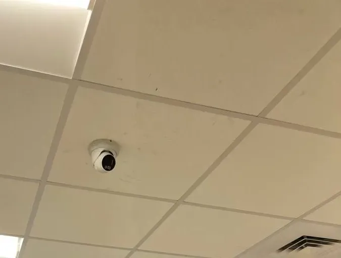 White turret camera on a ceiling tile near a light panel — example photo for Orange County classroom wings Orange County school security camera installation example photo: a white turret camera mounted on a beige ceiling tile next to a recessed light panel and an air vent