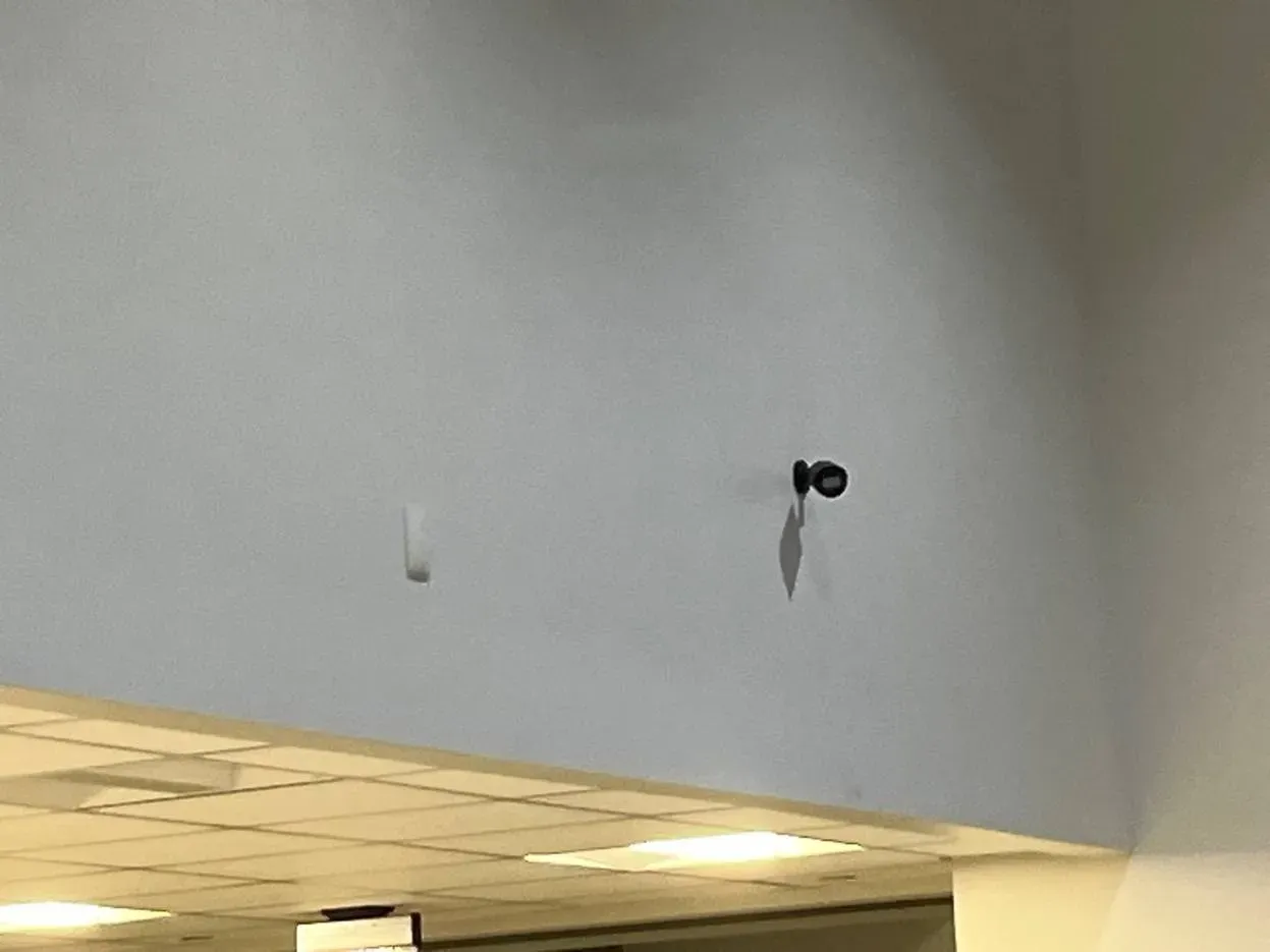 Black bullet camera high on a pale blue wall — Ulster County SmartPSS setup example Ulster County SmartPSS setup example photo: a small black bullet camera on a short bracket high on a pale blue wall above a drop ceiling edge, casting a shadow below it