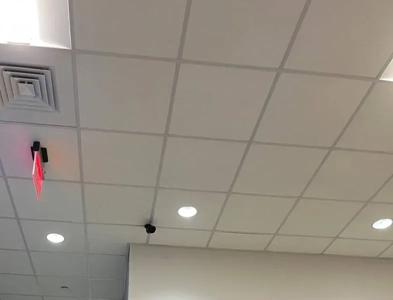Small ceiling camera near an exit sign — Ulster County SmartPSS support example Ulster County SmartPSS support example photo: a small dark camera on a white drop-ceiling grid between recessed lights, near a lit red exit sign and a square air vent