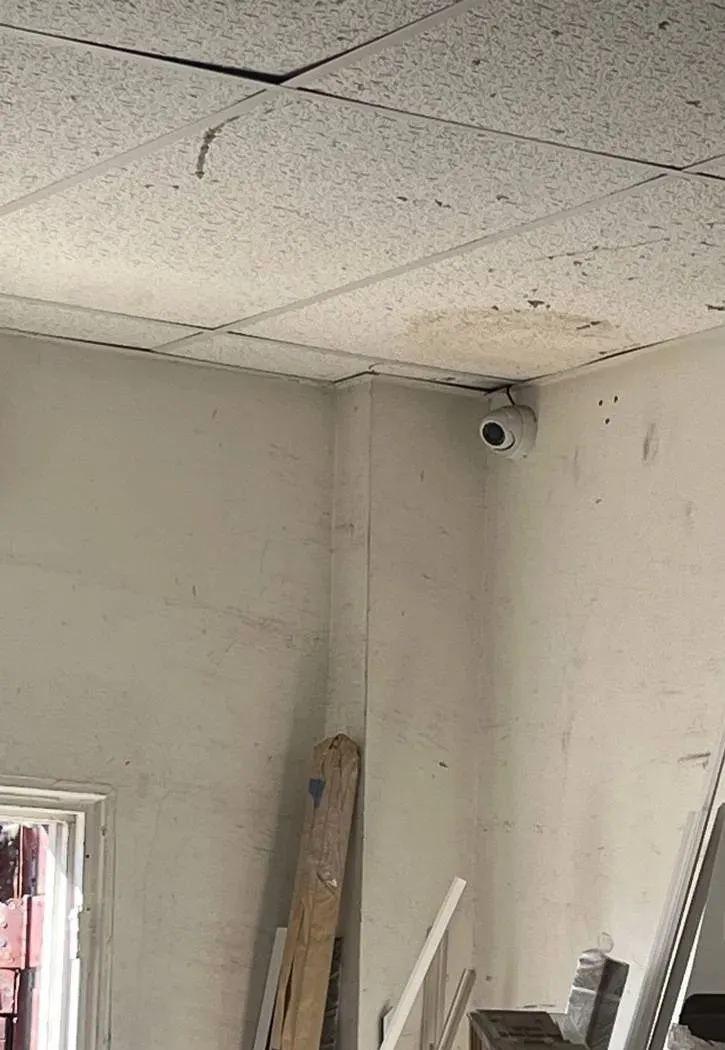 White turret camera in the corner of an unfinished room — Ulster County SmartPSS setup example Ulster County SmartPSS setup example photo: a white turret camera mounted in the corner of an unfinished room under stained ceiling tiles, with lumber leaning against the wall