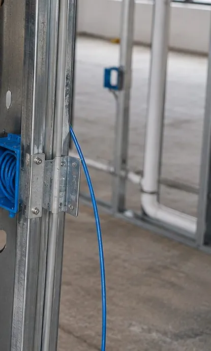 Sonos installation Battery Park City, Manhattan: a blue low-voltage box and blue cable fixed to a galvanised steel stud in open framing, a copper pipe stub and tall windows in the unfinished room behind
