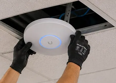 Sonos installation Battery Park City, Manhattan: black-gloved hands pressing a round white wireless access point with a blue ring light against an opened ceiling tile