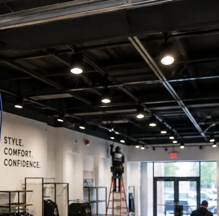 Sonos installation Battery Park City, Manhattan: a clothing shop floor under an open black ceiling with rows of round pendant and track lights, display racks and a worker on a ladder near the glass entrance