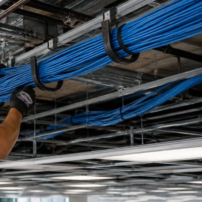 Cable bundles supported in J-hooks above a ceiling grid — Country Club, Bronx Sonos installation Sonos installation Country Club, Bronx: thick bundles of blue cable carried in black J-hooks above an open ceiling grid, with a gloved hand guiding the bundle into place