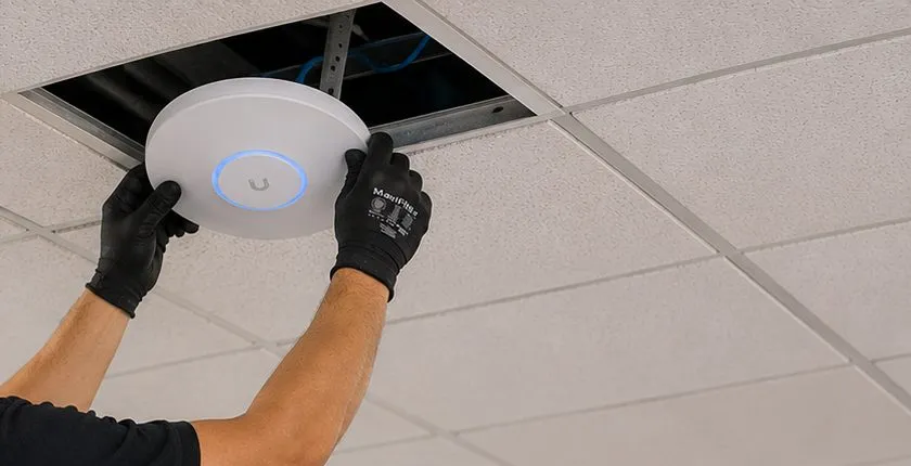 Wireless access point raised into an open ceiling tile — Country Club, Bronx Sonos installation Sonos installation Country Club, Bronx: gloved hands lifting a round white wireless access point with a glowing blue ring up into an open ceiling tile