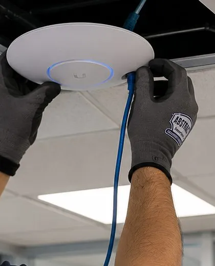 Wireless access point pressed into an open ceiling — Ditmas Park, Brooklyn Sonos installation Sonos installation Ditmas Park, Brooklyn: two gloved hands pressing a round white wireless access point with a glowing blue ring into an open ceiling, blue cable hanging below