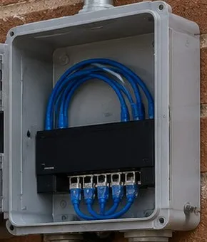 Open weatherproof box with patch cables on brick — Greenwich Village, Manhattan Sonos installation Sonos installation Greenwich Village, Manhattan: an open grey weatherproof box on a brick wall with loops of blue patch cable feeding a small black network switch inside