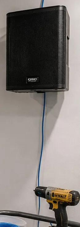 Wall-mounted box speaker with its cable drop — Greenwich Village, Manhattan Sonos installation Sonos installation Greenwich Village, Manhattan: a black box speaker fixed high on a pale wall with a blue cable running straight down from it to a yellow cordless drill below
