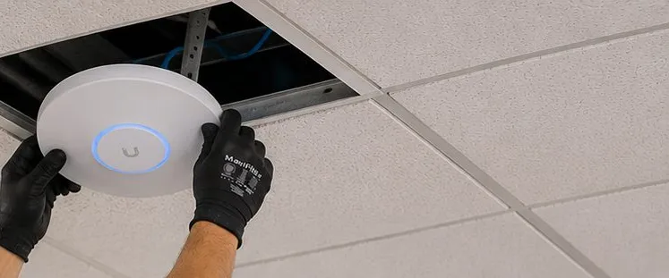 Access point pressed into a ceiling tile โ Grymes Hill, Staten Island Sonos installation Sonos installation Grymes Hill, Staten Island: two gloved hands pressing a round white wireless access point with a blue light ring into an open suspended-ceiling tile