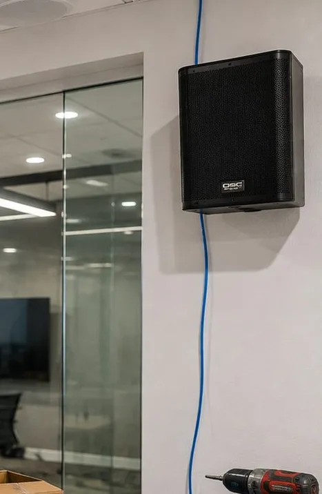 Wall speaker beside a glass partition โ Grymes Hill, Staten Island Sonos installation Sonos installation Grymes Hill, Staten Island: black box speaker mounted high on a white wall with blue cable running down beside it and a glass-walled room to the left