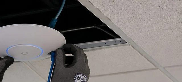 Sonos installation Long Island City, Queens: gloved hands pressing a white round wireless access point to a ceiling tile as a blue network cable rises into the gap