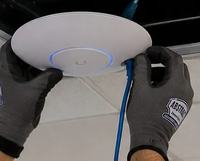 Access point raised to an open ceiling tile — Riverdale, Bronx Sonos installation Sonos installation Riverdale, Bronx: gloved hands fitting a white access point with a blue ring under an open ceiling tile