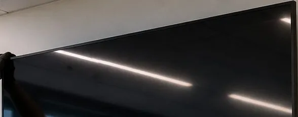 Top edge of a large screen being hung — Riverdale, Bronx Sonos installation Sonos installation Riverdale, Bronx: top edge of a large dark screen with light reflections, a gloved hand at one corner