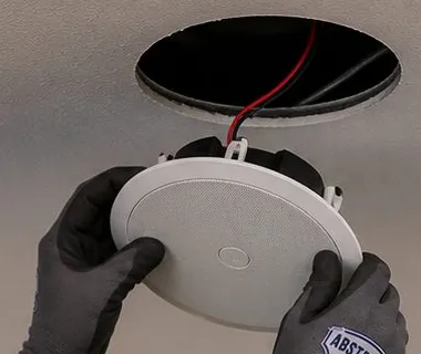 Ceiling speaker lifted to a round opening — Riverdale, Bronx Sonos installation Sonos installation Riverdale, Bronx: gloved hands lifting a round ceiling speaker to a cut opening with wire hanging out