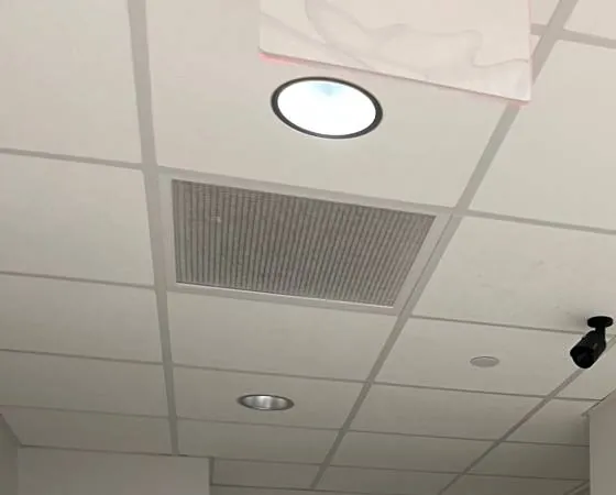 Perforated grille and round downlight in a tile ceiling — reference photo, Sonos installation St. George, Staten Island St. George, Staten Island Sonos installation reference photo: rectangular perforated grille set into a tile ceiling next to a round downlight and a small black camera on the grid