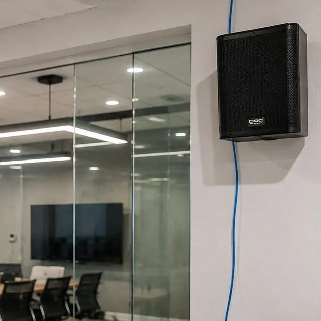 Sonos installation Upper East Side, Manhattan: black box speaker mounted high on a white wall with a blue cable running down from it, beside a glass-walled meeting room with a screen and chairs